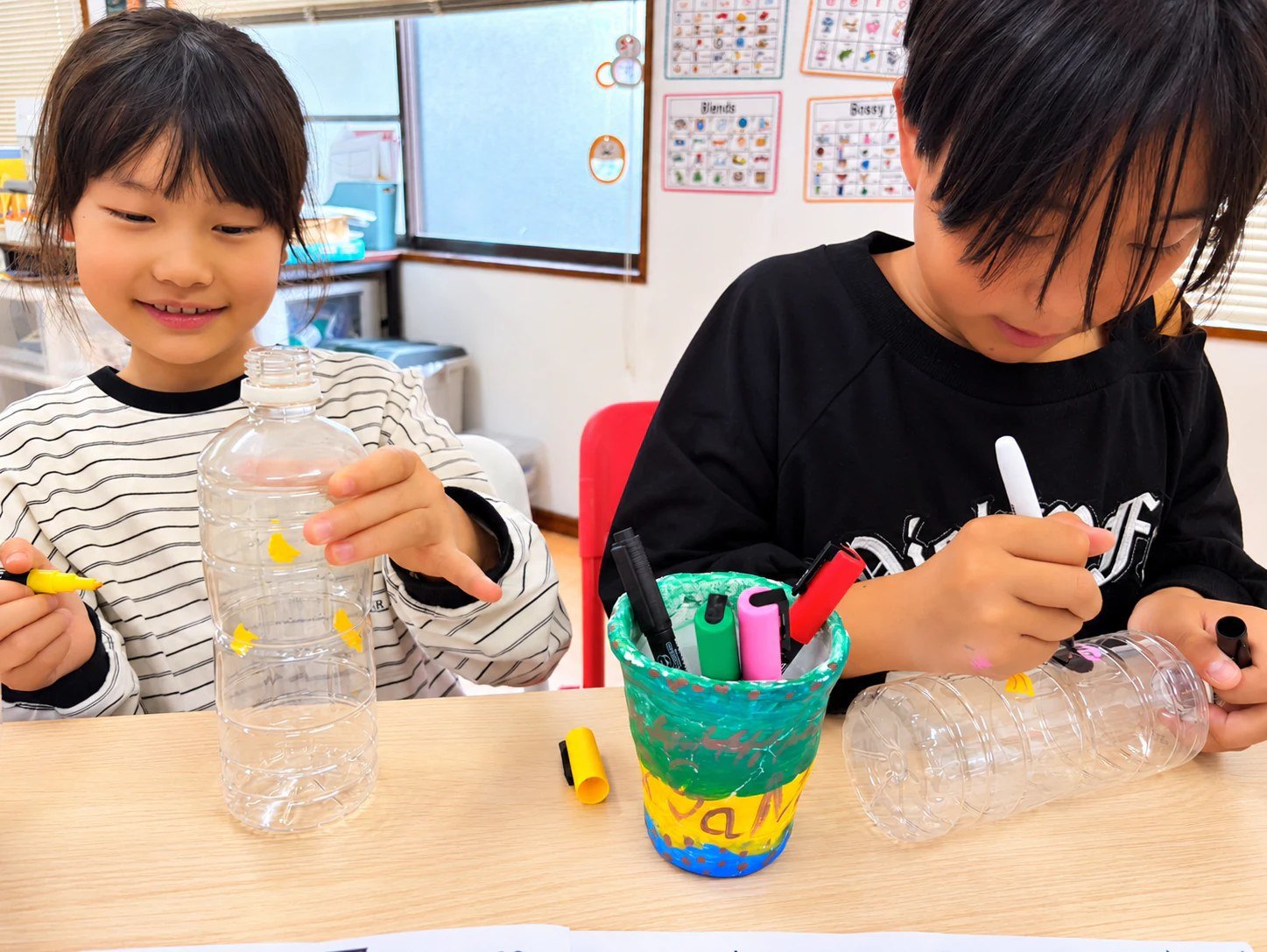 Children drawing on clear plastic bottles with colorful markers.