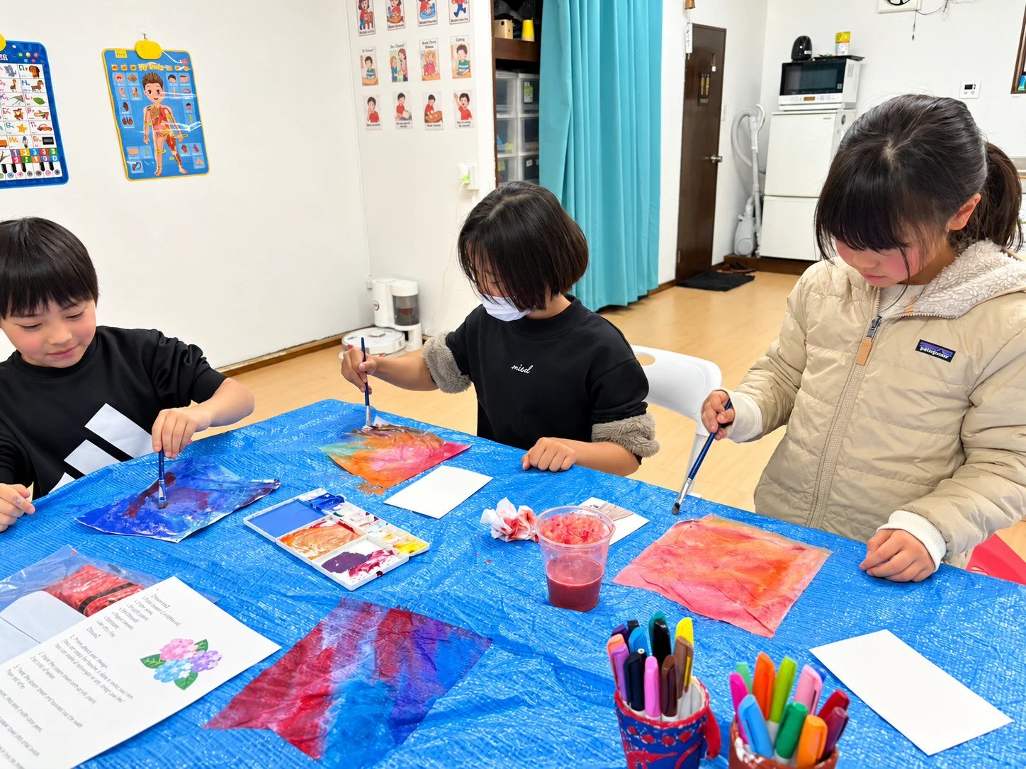 Children painting colorful sheets with brushes on a blue-covered table.