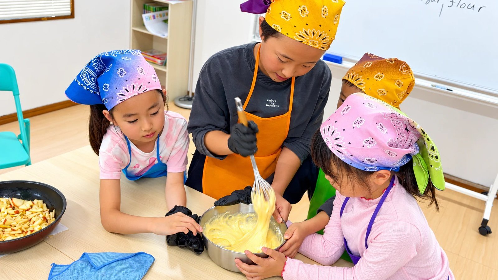 Children in headscarves and aprons mixing batter in a bowl with a teacher.