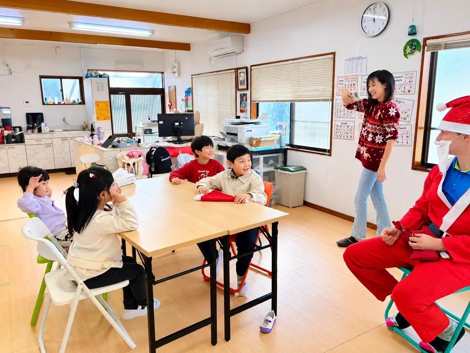 Children seated around classroom tables beside an adult in a Santa costume.