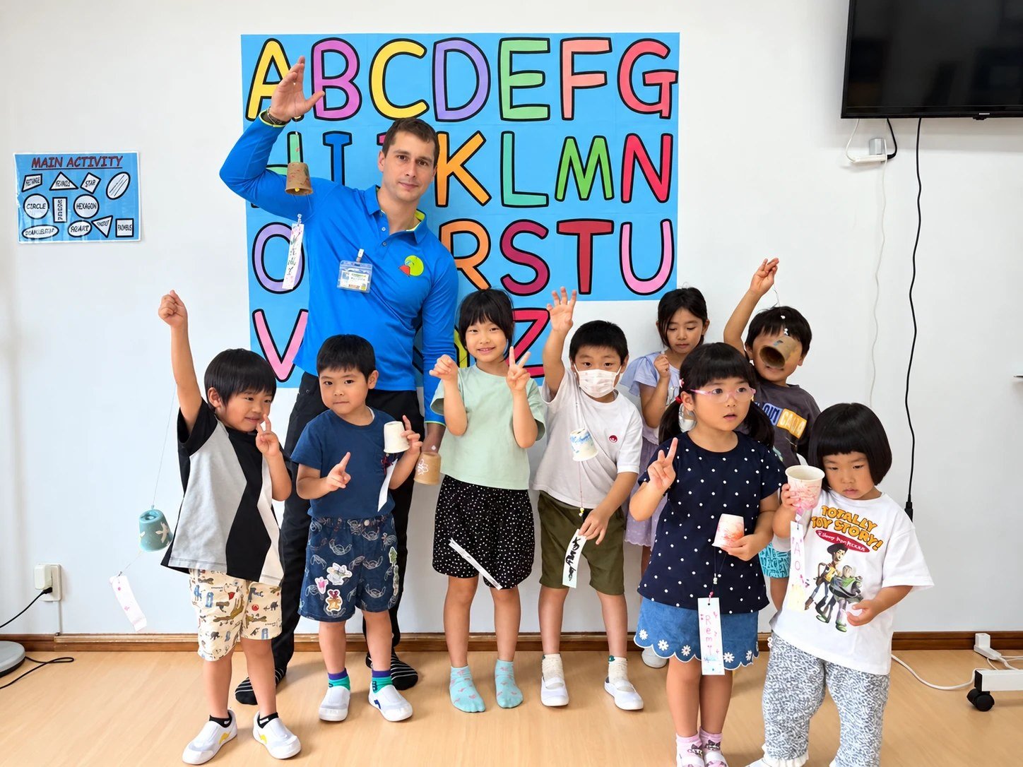 A teacher and children with paper cups in front of a colorful alphabet display.