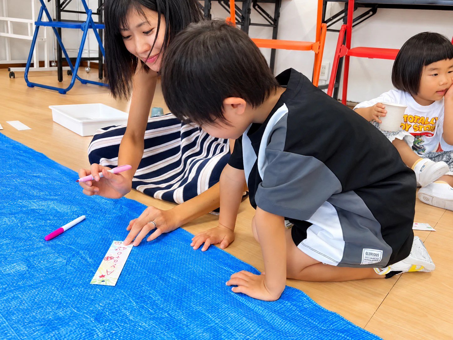 A teacher and child writing on a narrow strip of paper beside a blue sheet.