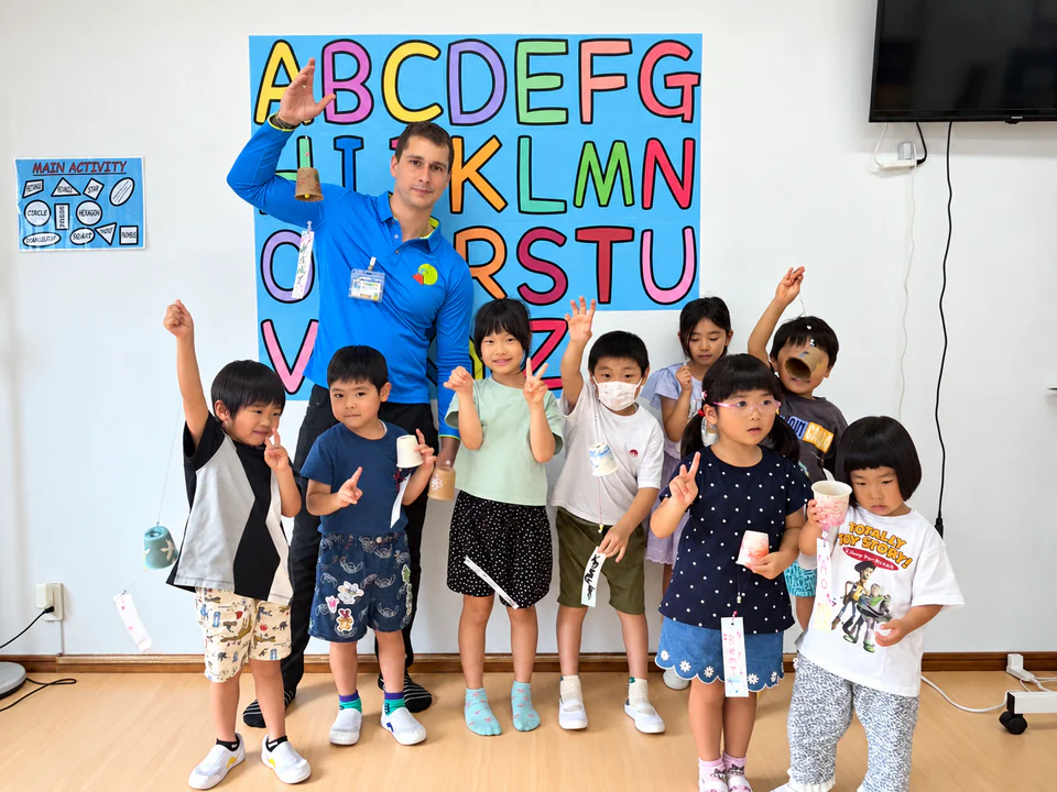 手作りの風鈴を持ち、アルファベットの掲示の前に集まるSELSの子どもたちと先生